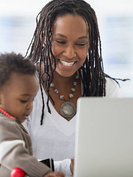 A single mother is answering emails and working on her website for her new start up company. She is watching her toddler son while working.